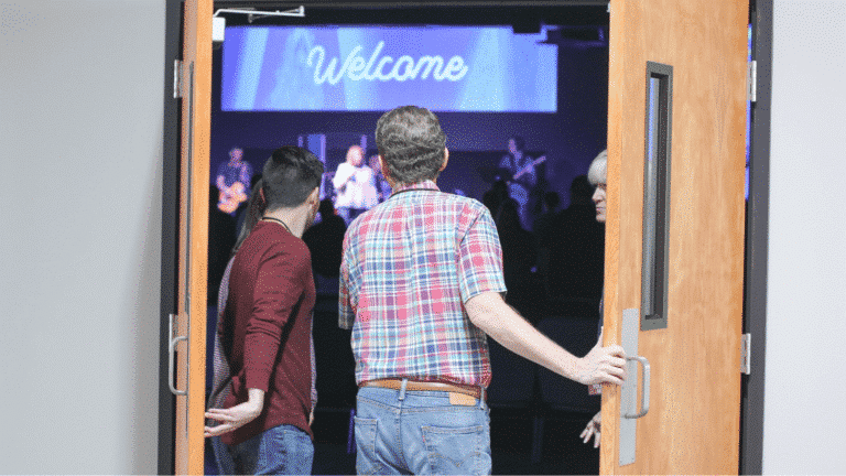 Two men standing at the back of church with the door open looking inside at the stage with a welcome sign. My Church Staff Guest Services