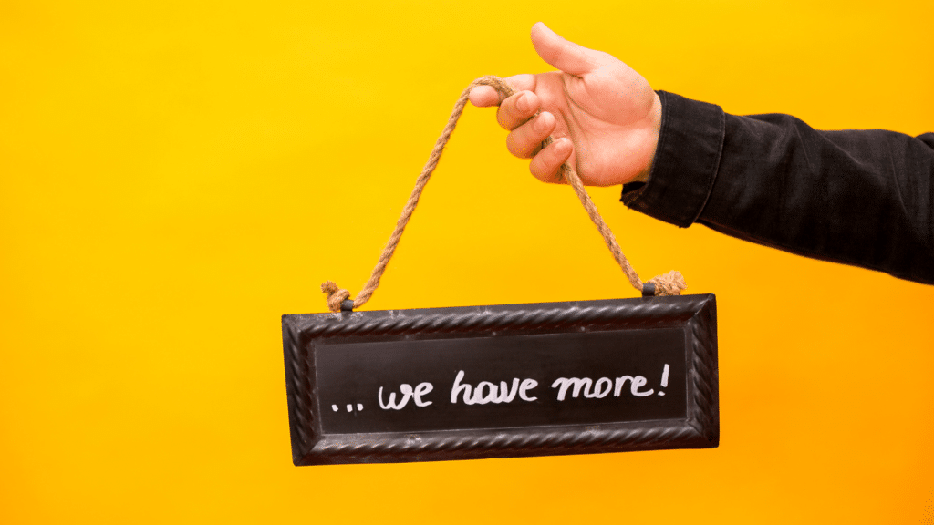 Yellow background. Black rectangle chalk sign that says....we have more! Hand holding the sign with a rope attached to it. Church Equip Curriculum